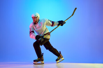 Man, hockey player wearing uniform and helmet, posing with stick against gradient blue background in neon light. Concept of professional sport, competition, game, tournament
