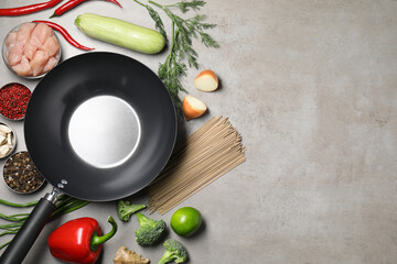 Empty iron wok surrounded by raw ingredients on grey table, flat lay. Space for text