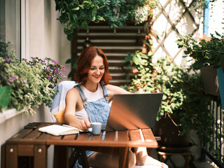A young woman with red hair enjoys working on her laptop surrounded by lush potted plants on a sunny balcony