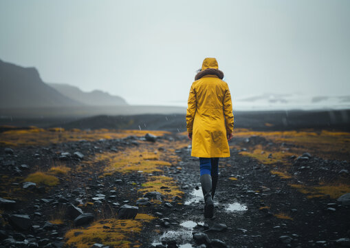 A woman in a yellow raincoat walks through a puddle under the cloudy sky - Powered by Adobe