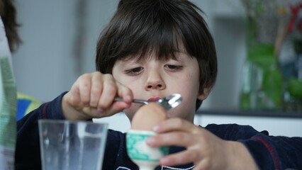 Child cracking open egg with spoon at breakfast table. One concentrated small boy opening...