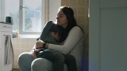 Mother holding child in arms leaning on kitchen wall resting together with eyes closed. Candid...
