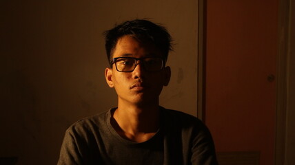A contemplative young man captured in dramatic lighting, sitting alone in a dim room. The photo...