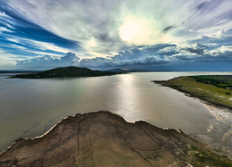 An aerial view of a tranquil lake bordered by lush green landscapes and rugged terrains, under a dramatic sky with sunlight piercing through the clouds. Chonlasit Dam, Saraburi, Thailand.