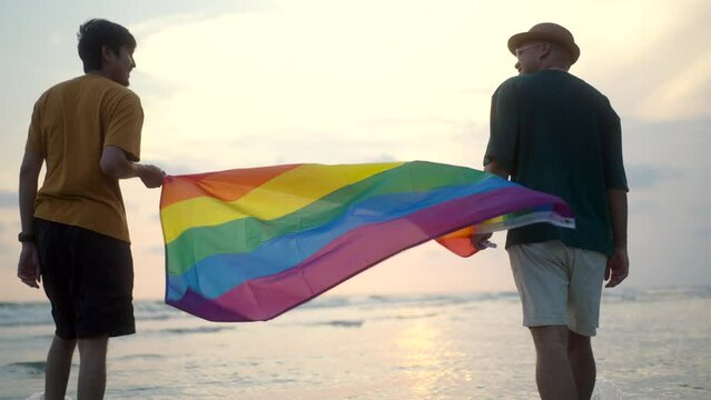 Romantic moment of bisexual gay male couple walking on the beach with their arms crossed with rainbow flag. LGBTQ man couple have a romantic moment on the beach. LGBT and homosexual concept.