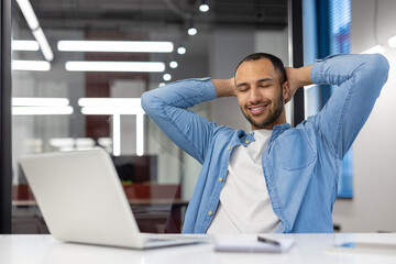 Smiling and relaxed young hispanic man sitting in the office at the desk, resting his hands behind his head and closing his eyes