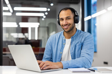 Portrait of a smiling young man sitting in the office with a laptop in headphones and looking confidently into the camera