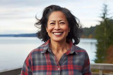 Portrait of a grinning asian woman in her 50s wearing a comfy flannel shirt isolated on serene lakeside view - obrazy, fototapety, plakaty