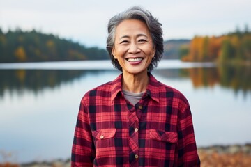 Portrait of a grinning asian woman in her 50s wearing a comfy flannel shirt over serene lakeside view - obrazy, fototapety, plakaty