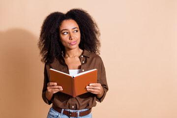 Portrait of minded woman with wavy hairstyle dressed brown shirt hold book look at logo empty space isolated on beige color background