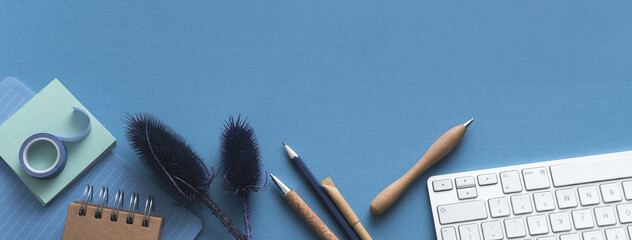 Workspace blue table with keyboard, wooden objects, tape, pencils and plant. Overhead view.