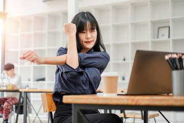 Office worker stretches at her desk in a bright office space, with a laptop open and a coffee cup...