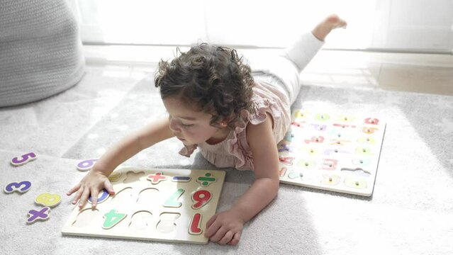 4K slow motion horizontal video of a smiling girl with curly hair lying on the floor playing with wooden puzzles of pieces and numbers. montessori materials. concept development and learning in the ch