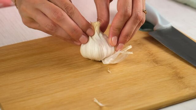 Woman cooking peeling garlic using knife on wooden board, hands close-up. Preparing food, meal on kitchen at home. Cooking dish. Culinary, cuisine, cook with fresh products, ingredients concept.