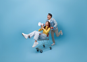 Asian girl sitting inside of shopping trolley and holding megaphone and Asian man pushing shopping cart running to supermarket isolated on blue background.