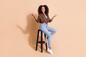 Full size photo of adorable woman dressed brown shirt sit on chair arms demonstrate objects empty space isolated on beige color background