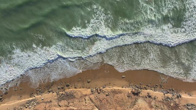 AERIAL TOP DOWN: Rolling ocean waves breaking and spilling along sandy shoreline. Mesmerizing view of waves gently lapping on the beach. Peaceful and empty stretch of Atlantic coastline in Portugal.