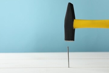Hammering metal nail on white wooden table against light blue background. Space for text