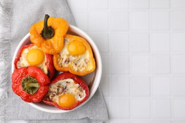 Delicious stuffed peppers with eggs in bowl on white tiled table, top view. Space for text