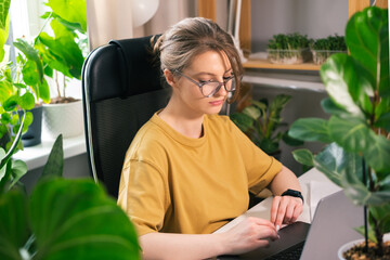Beautiful young woman working on laptop from home surrounded by indoor plants. Concept of remote...