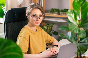 Beautiful young woman working on laptop from home surrounded by indoor plants. Concept of remote work, freelancing, online learning, in the urban jungle.