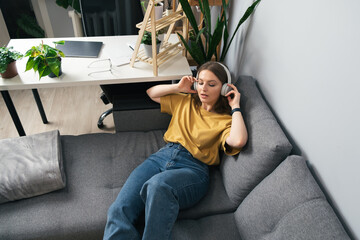 Young woman listening to music with headphones on the couch surrounded by indoor plants. Beautiful girl relaxing at home and listening to a podcast in a wireless headset.