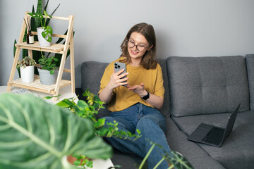 Girl talking on the phone while sitting on the sofa surrounded by indoor plants. Beautiful girl relaxing at home and looking at her phone. Jungle concept at home, remote work, freelancing