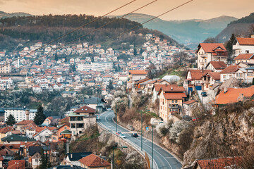 Naklejka premium At sunset, Sarajevo's cityscape unfolds, with winding roads weaving through colorful neighborhoods against the backdrop of majestic mountains.