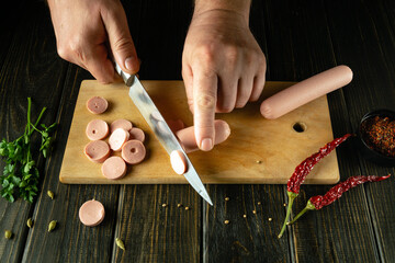 The chef uses a knife to cut a sausage on a kitchen board before frying. Low key concept of preparing a diet dinner on the kitchen table