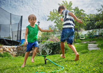 Two happy boys play and jump around sprinkler in sunny garden