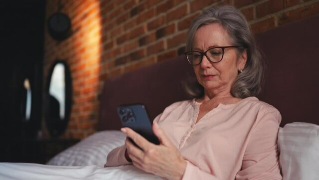 Happy woman in her 60s lying in bed and scrolling social network on smartphone