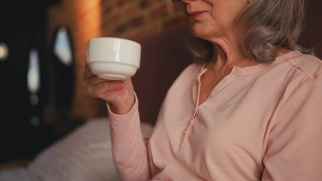 Happy woman in her 60s enjoying coffee in bed, morning ritual, leisure time
