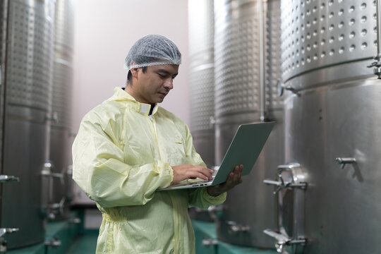 Winemaker inspecting quality the process of fermentation during manufacturing in winery factory and stainless tank. Male winemaker working with laptop computer in winery factory