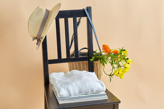home decor and design concept - flowers in wicker bag or basket, clothes, hat and magazines on vintage chair over beige background