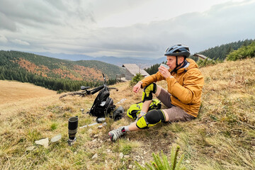 Naklejka premium Man cyclist drinking a cup with tea, wearing bike helmet, sitting on grass in the mountains, resting after riding electric bike. Mountain e-bike lying on the ground next to him.