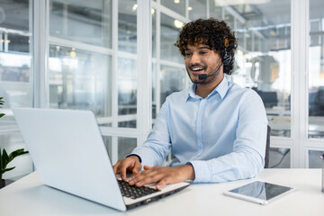 Cheerful male customer service representative with headset working on laptop in a bright, contemporary office setting. Professional communication and support.