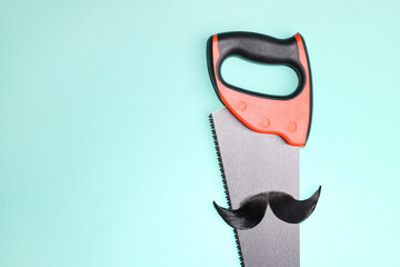 Man's face made of artificial mustache and hand saw on light blue background, top view. Space for...