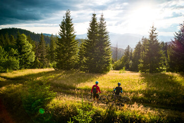 Two cyclists men riding electric bikes outdoors at sunset. Back view of male tourists resting on the hill, enjoying beautiful mountain landscape in the evening. Concept of active leisure.
