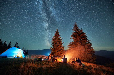 Night camping in mountains under starry sky. Group of people tourists having a rest near campsite, burning campfire and illuminated tent. Concept of tourism, hiking and adventure.