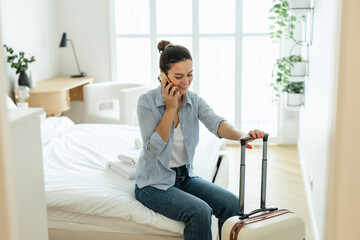 Young woman with a suitcase sitting on bed in hotel room and using phone
