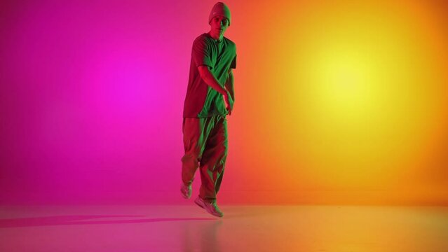 Young athlete man, dressed streetwear dancing freestyle moves in motion in neon light against gradient pink-yellow background. Concept of art, hobby, sport, creativity, fashion and style, action. Ad