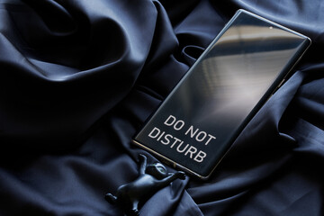 Modern smartphone - a cell phone with do not disturb inscription on the screen and a black toy kitten on a dark silk fabric background. Concept of rest and solitude of an introvert - obrazy, fototapety, plakaty