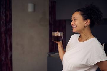 Multi ethnic curly haired pretty woman in white t-shirt of her pajamas, holding a cup of freshly...