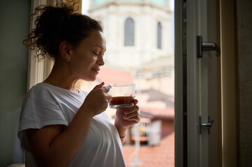 Happy woman with her eyes closed, holding a cup of freshly brewed coffee, standing by window...