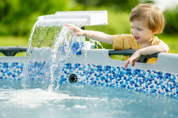Cute funny toddler boy having fun by an outdoor pool. Kid playing with water. Family fun in a pool. Summer activities for the entire family. - obrazy, fototapety, plakaty