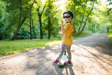 Adorable little boy riding his scooter in a city park on sunny summer evening. Young child riding a roller with a helmet on.