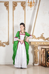 Beautiful smiling woman in green rococo style medieval dress standing near fireplace