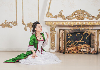 Beautiful smiling woman in green rococo style medieval dress sitting on the floor near fireplace