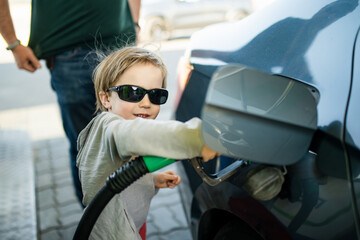 Cute little blond boy holding pump nozzle. Small kid helping father to fuel the car at a gas...