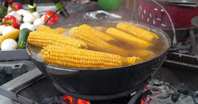 Boiled corn lies in big cauldron with hot water. Cooking over an open fire. Other vegetables lies nearby